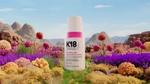 Bottle of K18 HeatBounce conditioning heat protectant displayed in a vibrant, colourful landscape with rocky mountains in the background and clusters of bright, spherical plants in shades of purple, pink, and yellow in the foreground.