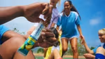 Close-up of a hand holding a Liquid I.V. Hydration Multiplier packet in lemon-lime flavour, with a group of people in sports uniforms gathered in the background on an outdoor field under a clear blue sky.