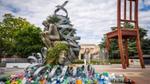 Male statue with his head in his hands sitting on a woman’s head, surrounded by metal coils, fresh flowers and waste plastic