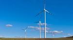 A row of wind turbines stand in a ploughed field, set against a bright blue sky