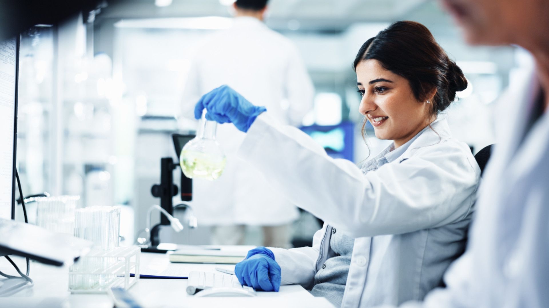 A scientist in a lab coat smiles while examining a flask of light green liquid at her workstation