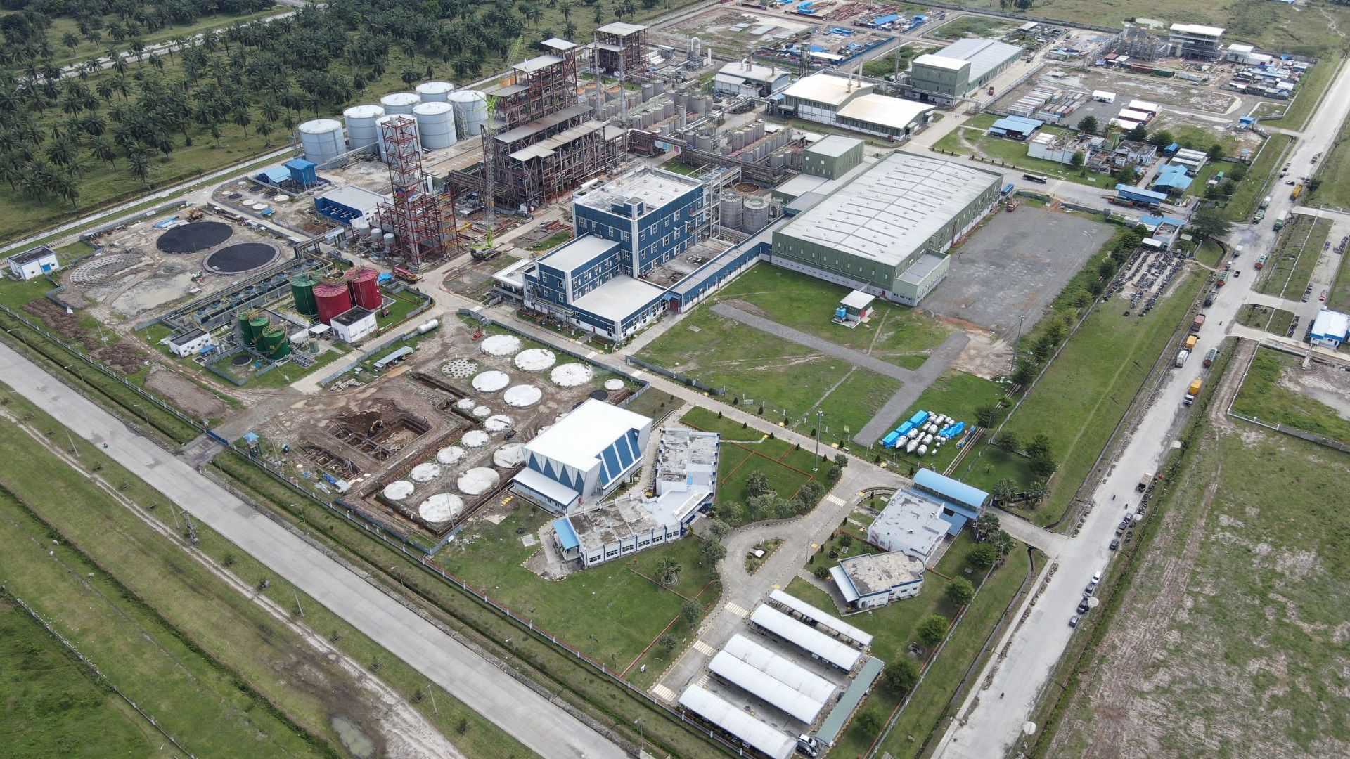 An aerial view of an industrial facility featuring buildings, large storage tanks and some green spaces, surrounded by roads and trees in the background.
