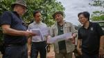 Four individuals are engaged in a conversation and looking at printed maps in a field/plantation setting. The group includes people dressed in casual and outdoor work attire, one wearing a wide-brimmed hat and another in a shirt with a WWF logo.
