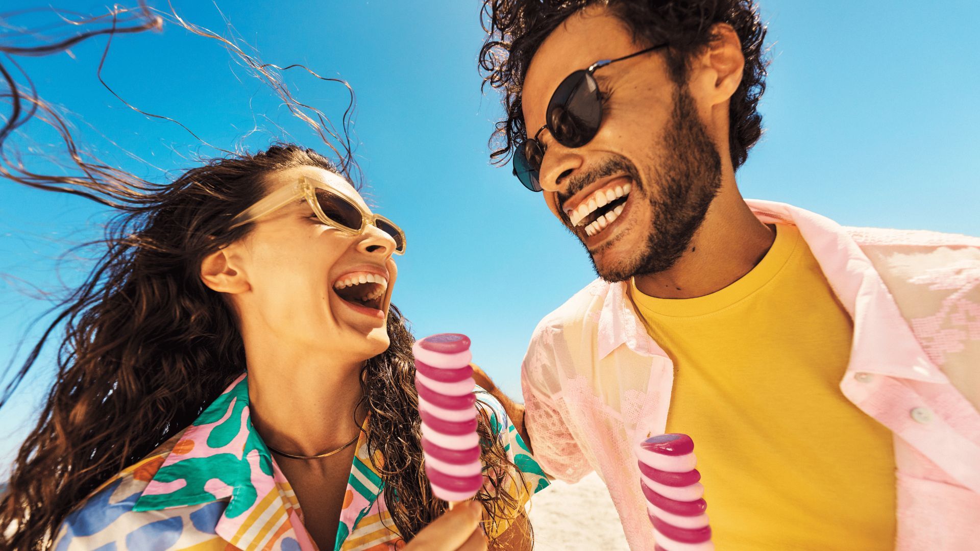 A woman and man, both with dark hair and sunglasses, are smiling at each other while each holds a Twister ice cream