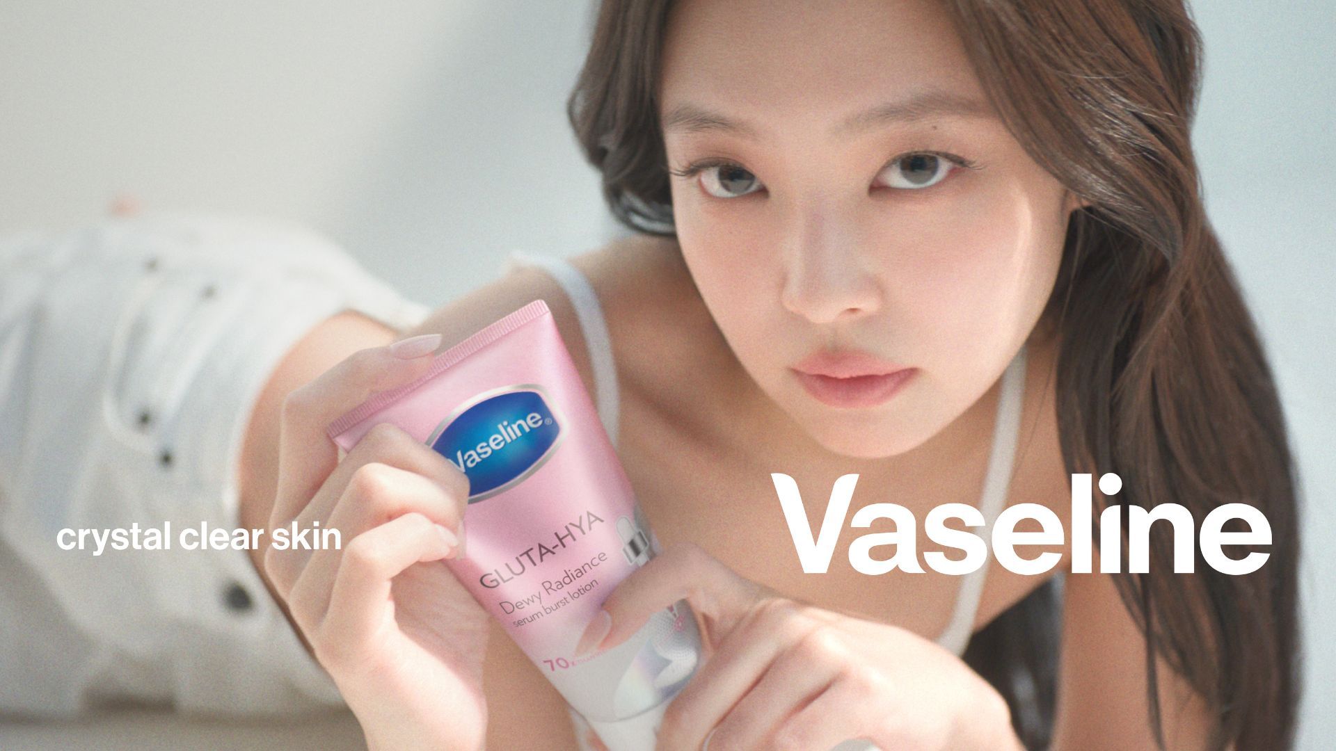 A person holds a pink tube of Vaseline Gluta‑Hya Dewy Radiance serum lotion close to the camera while looking forward, with soft lighting and a neutral background; on‑screen text reads “crystal clear skin” and “Vaseline”.
