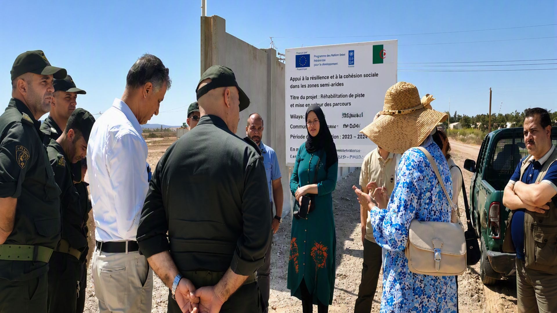 Officials and project staff stand outdoors discussing a development project beside an informational signboard.