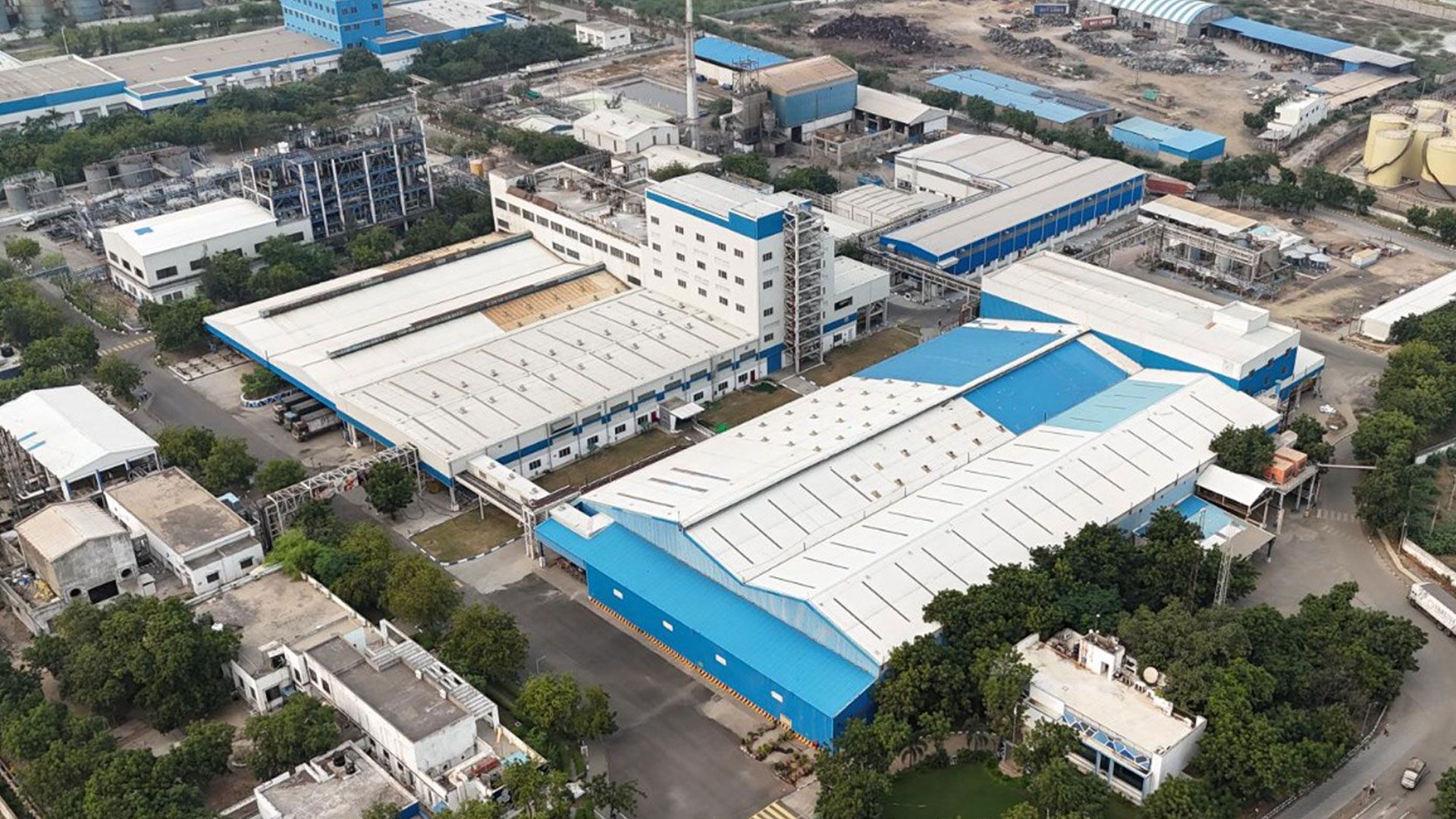 Aerial view of Unilever’s Gandhidham personal care site in India showing interconnected factory buildings with blue roofs, a central multi-storey processing unit with pipes and a chimney, loading bays, storage areas and access roads within a developed site.