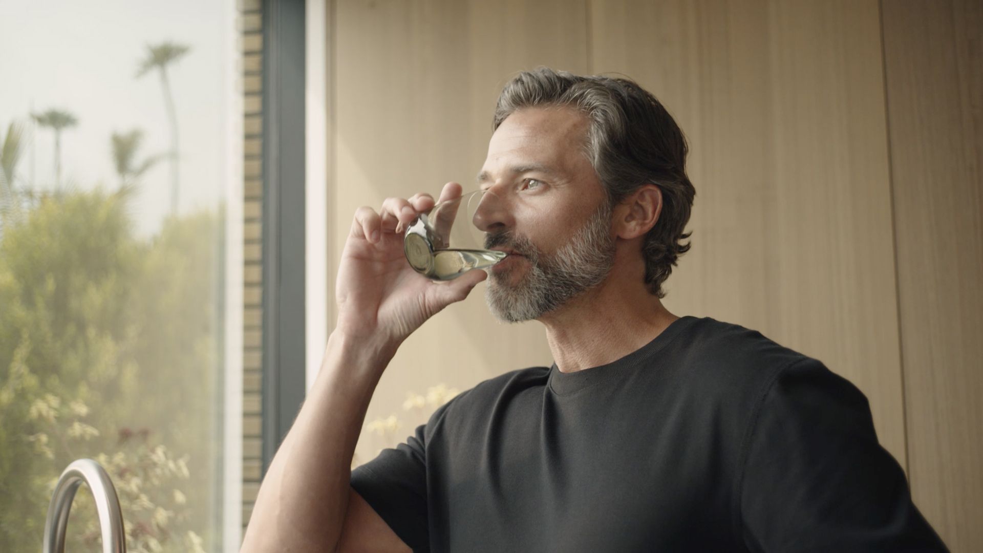 Person indoors holding a glass near their mouth, wearing a black shirt, with a blurred view of greenery outside the window.
