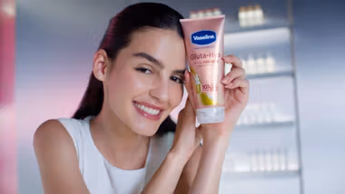 A white woman with dark hair and glowing skin, holding a tube of Vaseline Gluta Hya Dewy Radiance cream. Pink, premium packaging.