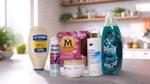 A variety of Unilever's Power Brands arranged on a kitchen counter. From left to right, you can see a bottle of Hellmann's Real Mayonnaise, a Degree deodorant spray labelled "WHOLE BODY DEO", a box of Magnum ice cream labelled "UTOPIA", a bottle of Dove body lotion pro-ceramides serum, a jar of Dove Intensive Repair cream and a bottle of Persil Wonder Wash. In the background, there's a modern kitchen with plants and various items on the shelves.