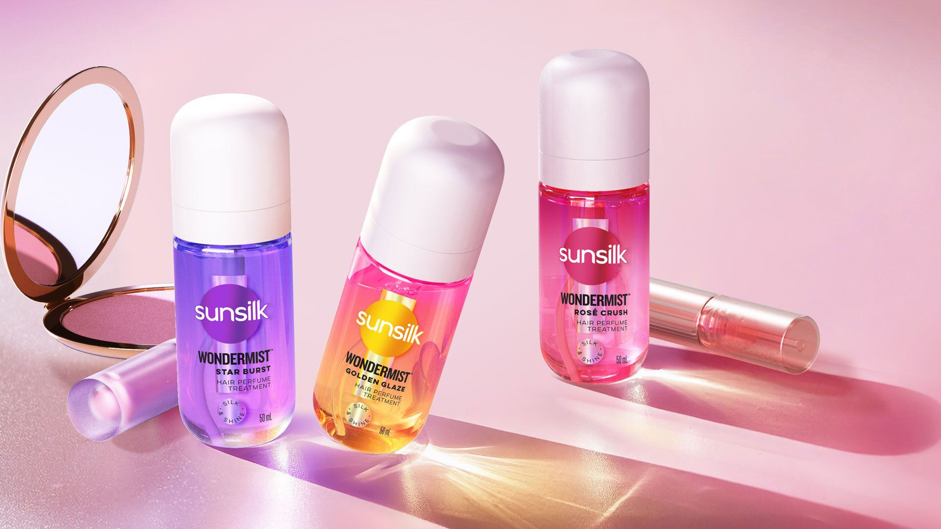 Three Sunsilk hair perfume bottles in vibrant colors on a pink backdrop, beside a compact mirror and a lip gloss.