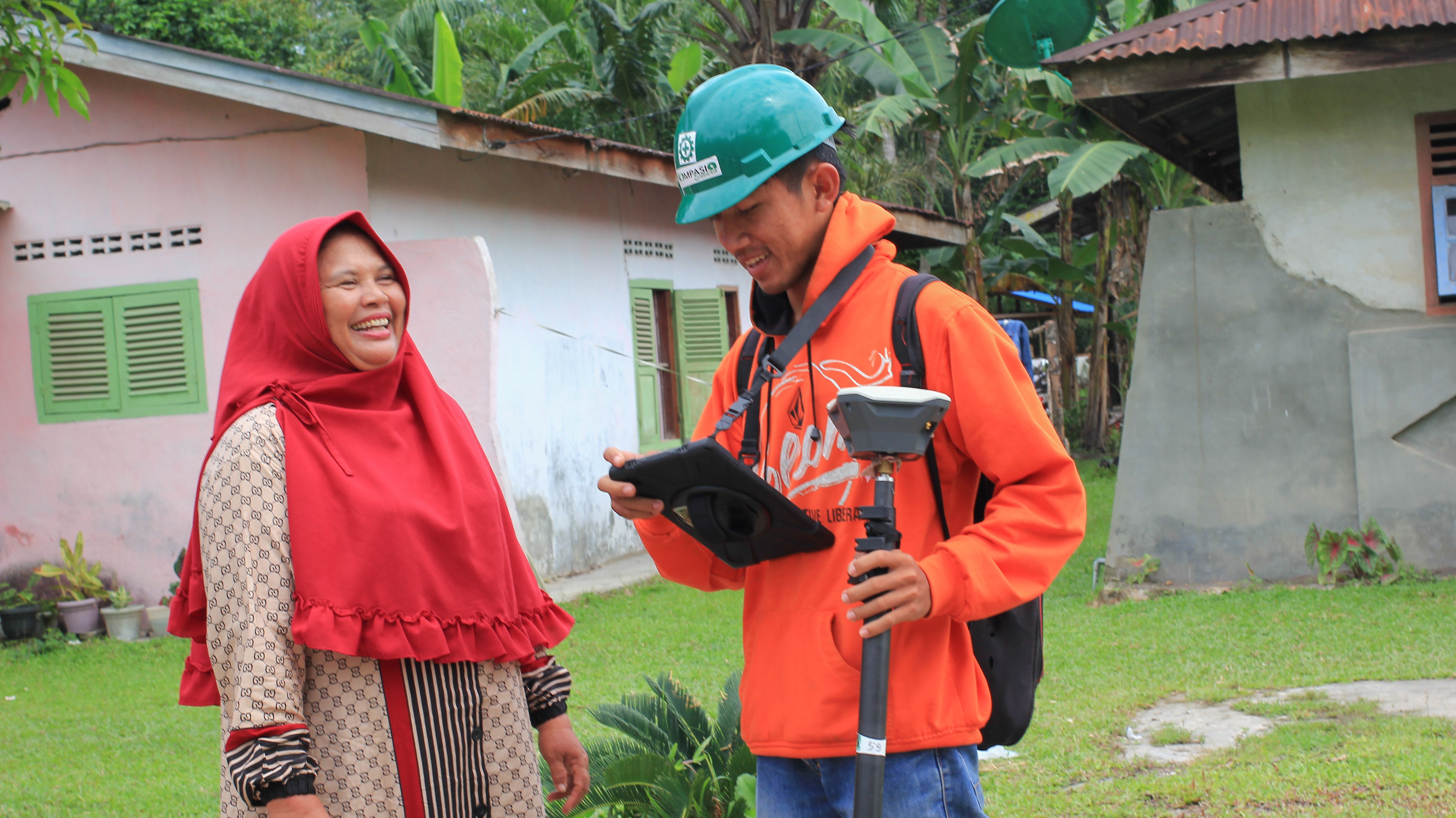 A man in an orange hoodie and green hard hat is holding surveying equipment and a tablet, smiling while engaging with a woman wearing a red headscarf. They are standing outdoors in front of a house with greenery in the background.