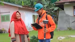 A man in an orange hoodie and green hard hat is holding surveying equipment and a tablet, smiling while engaging with a woman wearing a red headscarf. They are standing outdoors in front of a house with greenery in the background.