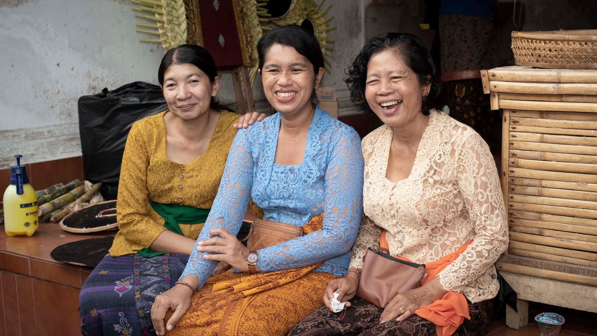 Three women in traditional clothing sit on a tiled step. They are surrounded by baskets, bamboo sticks, and household items in an outdoor setting.