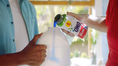 A close-up of a person pouring blue liquid detergent from an OMO-branded bottle into a larger white plastic container which a second person is holding.