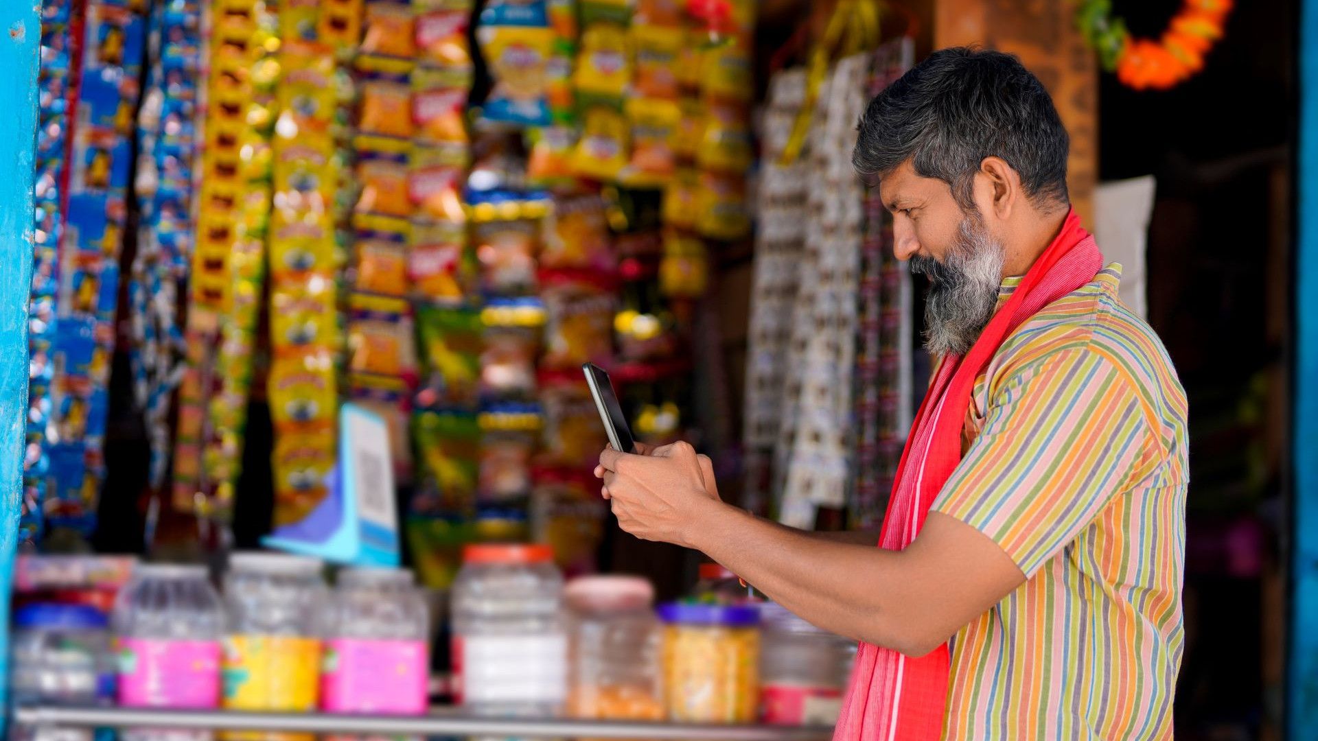 A shopkeeper wearing a striped shirt and a red scarf around his neck is using a smartphone while standing in front of a colourful shop display. The shop is filled with a variety of products, including packets hanging on the wall and jars of goods on the counter.