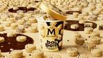 A tub of Magnum BonBon White Chocolate & Cookie ice cream with a gold lid, surrounded by small, round white chocolate and cookie bonbons scattered on a beige surface with dark chocolate patches.