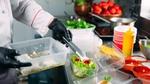 Chef in a commercial kitchen placing food into a takeaway container, with various bowls of ingredients on the work surface