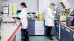 Two technicians in white lab coats and safety goggles working in a research lab, equipped with various devices, labelled drawers and containers.