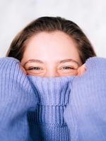A woman pulls her cosy sweater up over her face to below her glinting eyes.