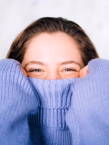 A woman pulls her cosy sweater up over her face to below her glinting eyes.
