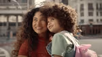 A woman holding a child with a pink backpack, with a city street in the background. They are both smiling.