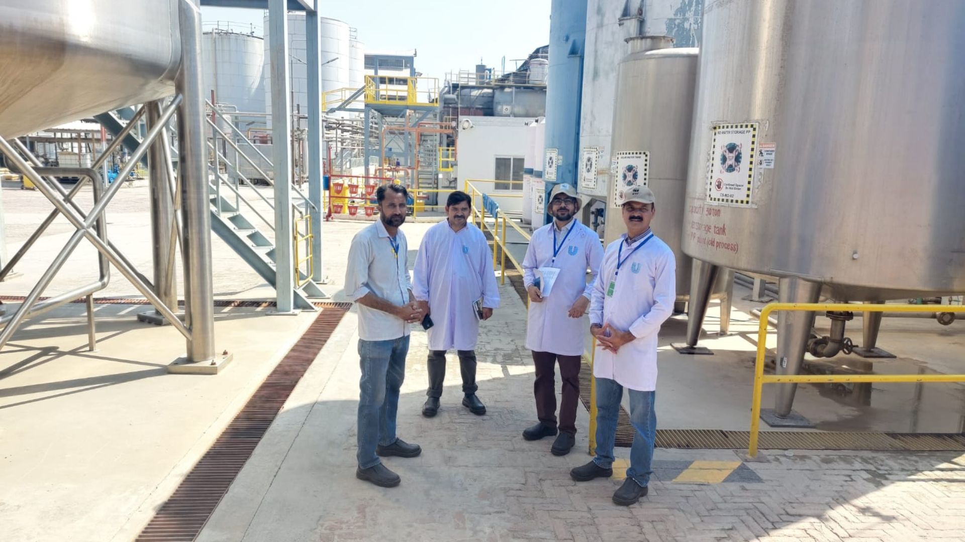 Four factory workers stand near large stainless steel tanks at an industrial processing plant