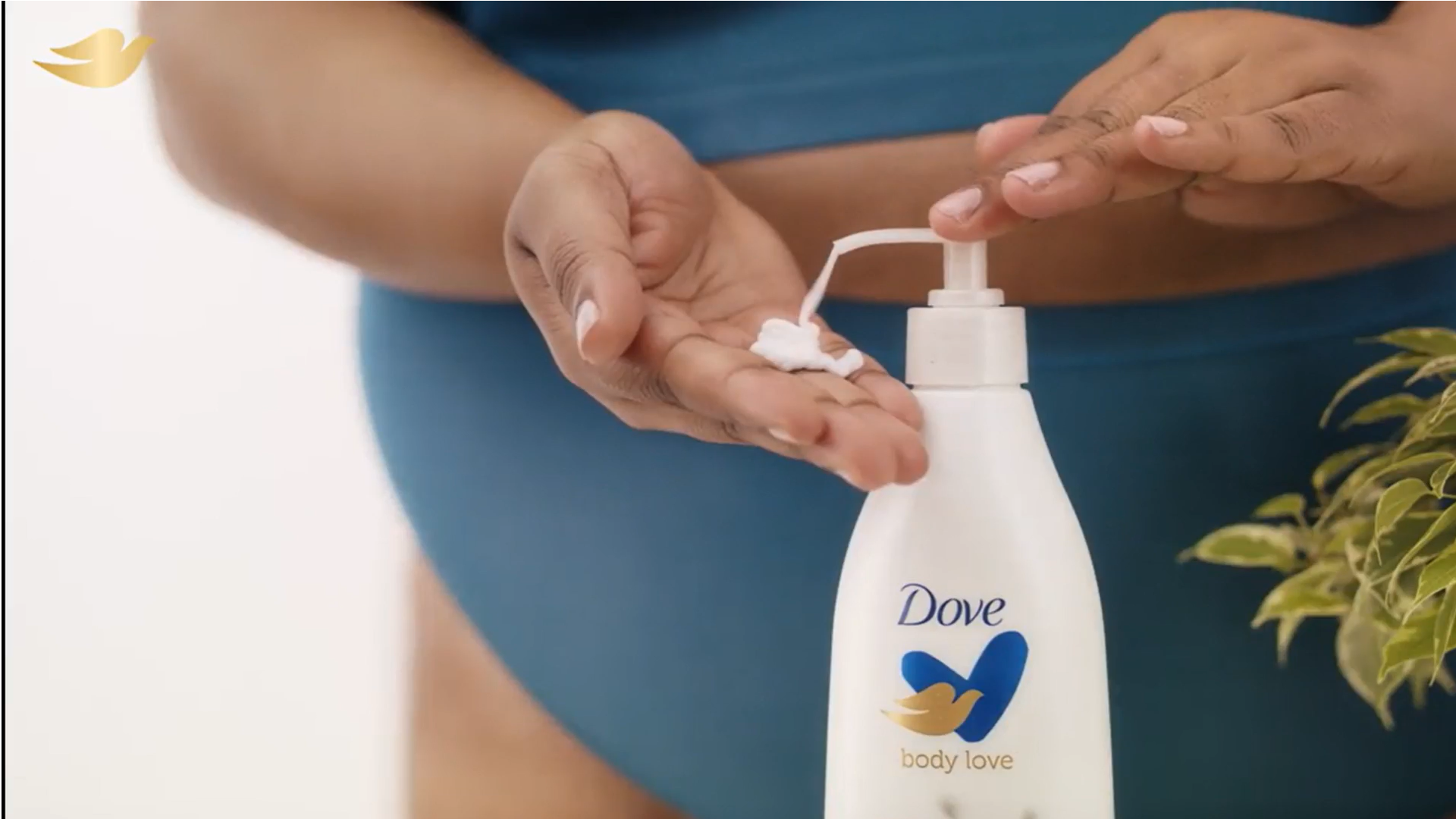 A woman in teal underwear pumps Dove Body Love lotion into her hand. Her face isn’t pictured. White bottle, Dove logo.