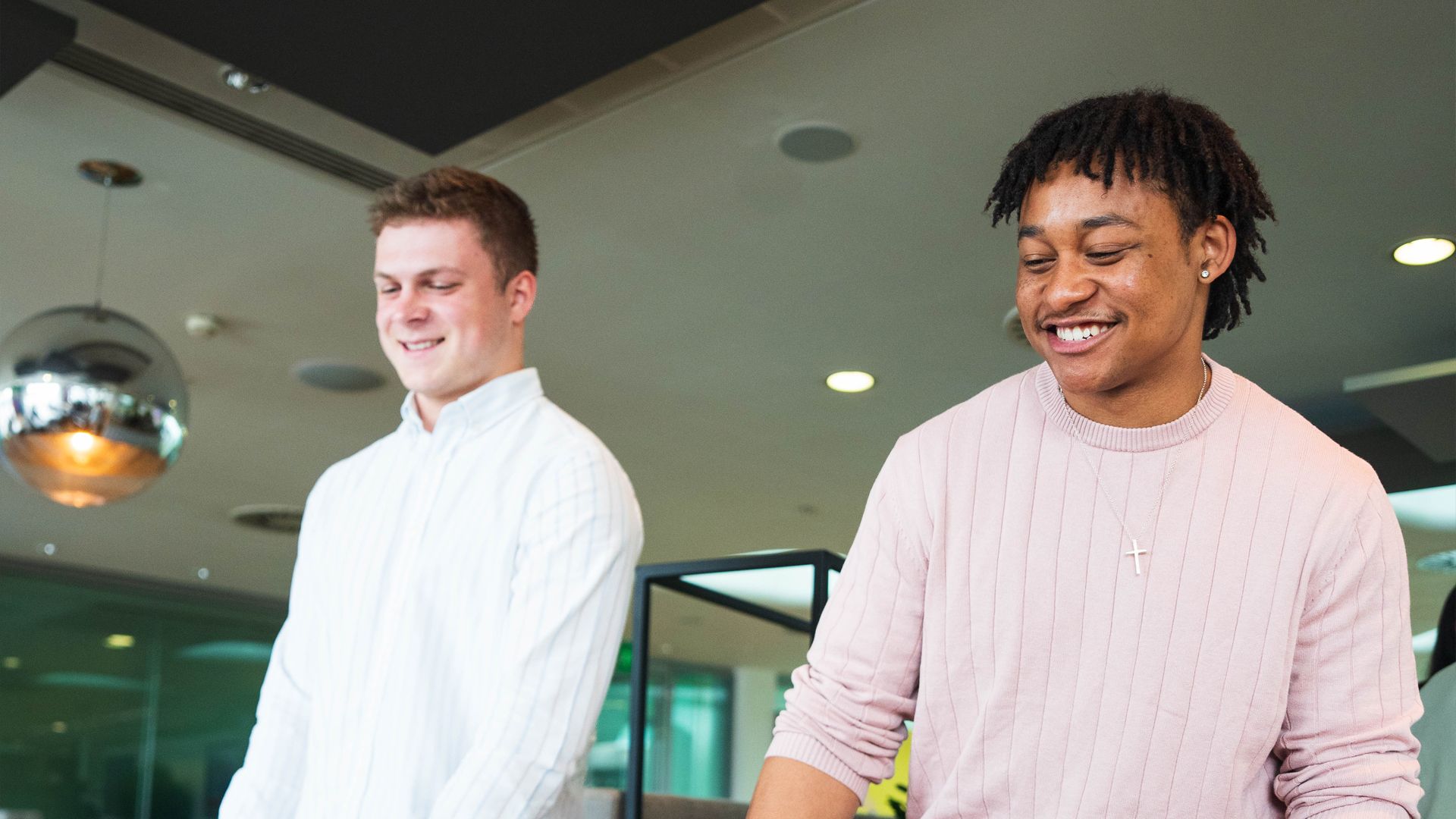 Two people standing in a modern office with stylish lighting and decor. One wears a white shirt, the other a light pink sweater and a cross necklace.