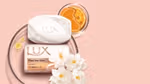 Two bars of LUX soap – one packaged, one not – from the Flaw-less Glow range are on the left of the image. Several jasmine flowers sit alongside the soap. This all sits on a pink beige background.