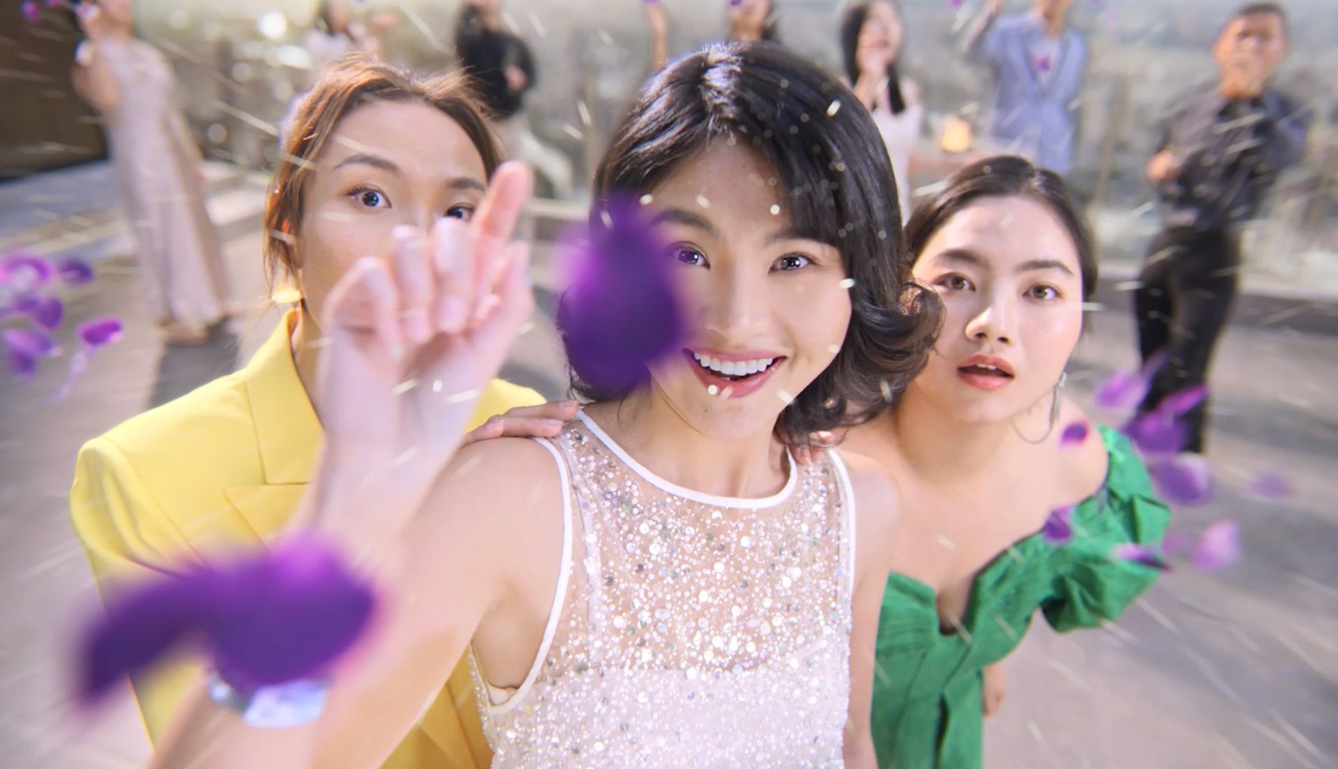 Group of people dressed in elegant outfits, including a sequined top and a green dress, surrounded by falling purple flower petals in an outdoor setting.