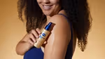 Woman with curly hair holding Vaseline Radiant X Replenishing Hydrating Body Oil spray. The product is on her arm.