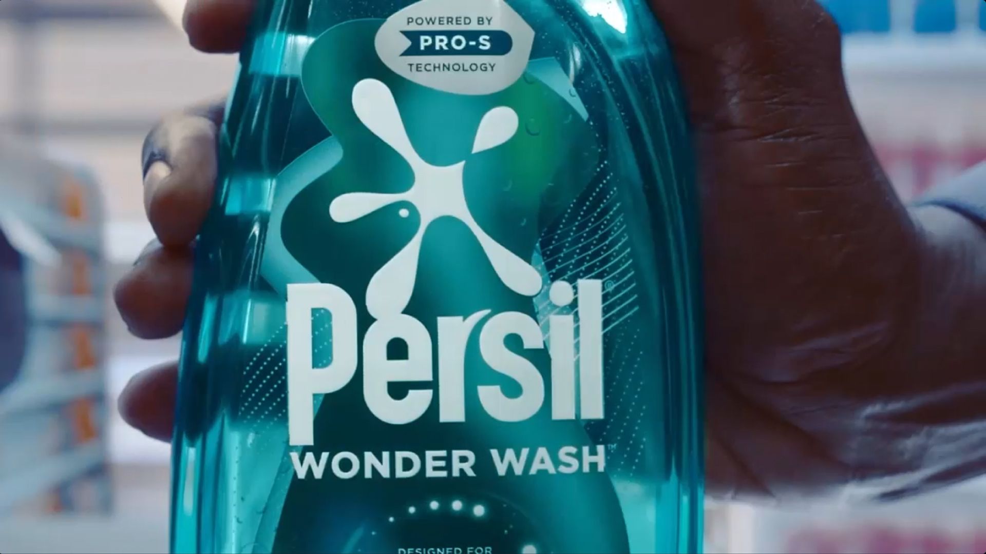 Close-up of a hand holding a green bottle of Persil Wonder Wash detergent, featuring the Persil logo, 'WONDER WASH' text, and a label reading 'POWERED BY PRO-S TECHNOLOGY'.