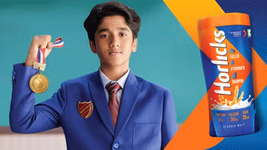 A schoolboy wearing a blue blazer with a ‘Head boy’ badge pinned to it stands with his right arm bent, fist clenched, grasping a 1st place medal. To the right of him is a jar of Horlicks.