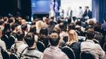 A speaker presenting on stage at a conference room. The view is from the back of the room showing a number of unrecognisable people in the audience