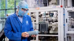 A person wearing a blue Unilever lab coat, hairnet, safety glasses and face mask examines a grey plastic bottle in a factory setting, with industrial equipment in the background.