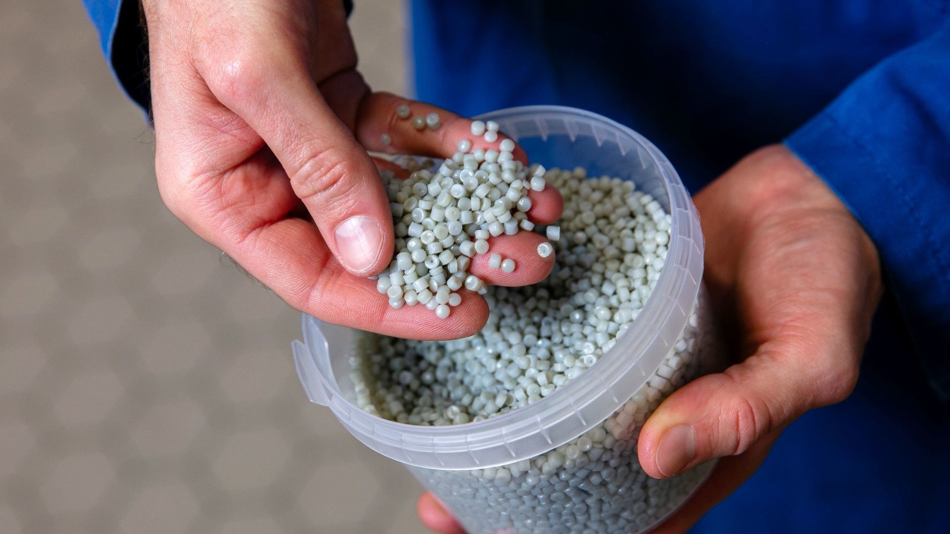 A close-up of hands holding small, grey plastic pellets. The pellets, which are all uniform in size and shape, are being scooped from a clear plastic container.