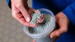 A close-up of hands holding small, grey plastic pellets. The pellets, which are all uniform in size and shape, are being scooped from a clear plastic container.