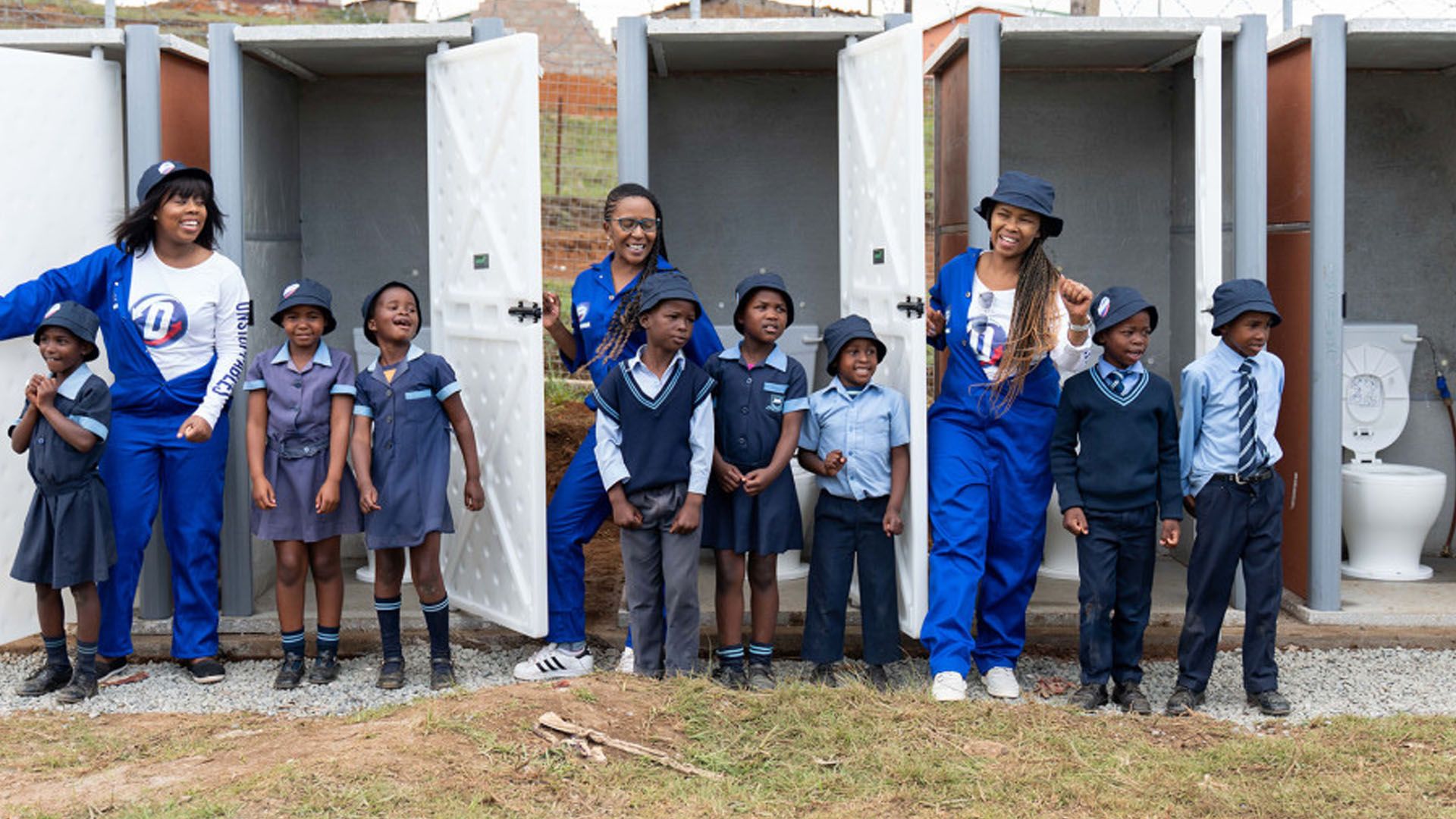 Three women in Domestos-branded blue overalls and several schoolchildren in uniform standing outside a row of toilets.