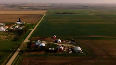 Aerial view of some rural farmland with several buildings including barns, surrounded by vast expanses of green and brown agricultural fields.