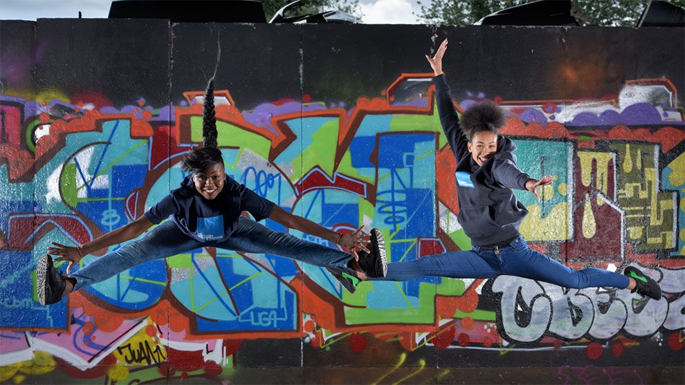 Two girls jumping in the air with their arms and legs outstretched in front of a wall covered with colourful graffiti.