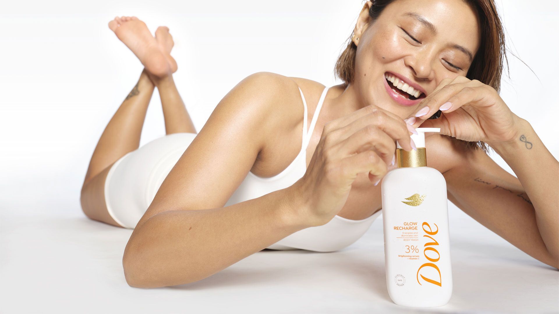 Person lying on their stomach in a white outfit, smiling while holding a bottle of Dove Glow Recharge body lotion with a pump dispenser.