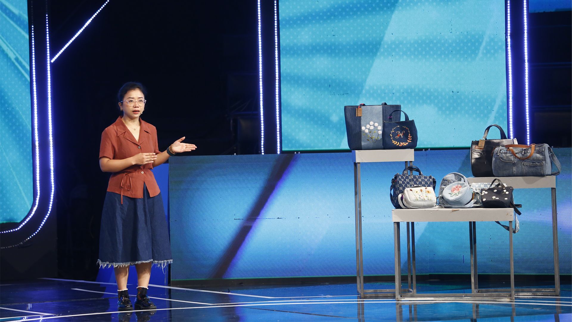 A woman speaking on stage, gesturing towards some display shelves. On the shelves are a range of bags of varying designs and sizes.