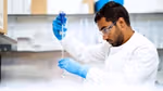 A person in a science lab wearing gloves and eye protection adding liquid to a test tube.
