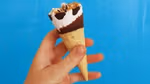 A hand holding a Cornetto ice cream in blue and white packaging. Topping is chocolate and nut. Set on a blue background.