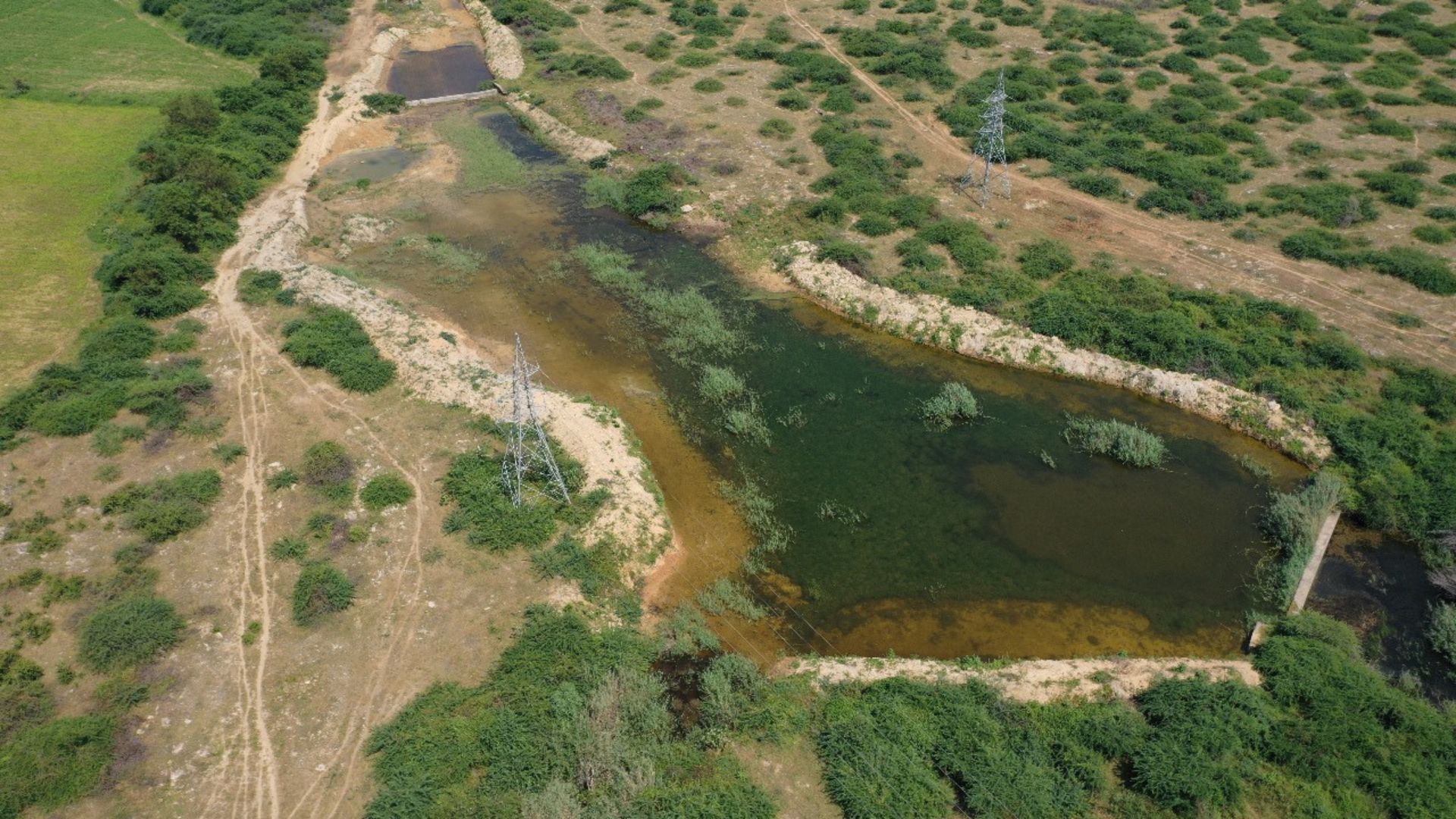 A narrow river with green vegetation flows through dry farmland crossed by metal power line towers.