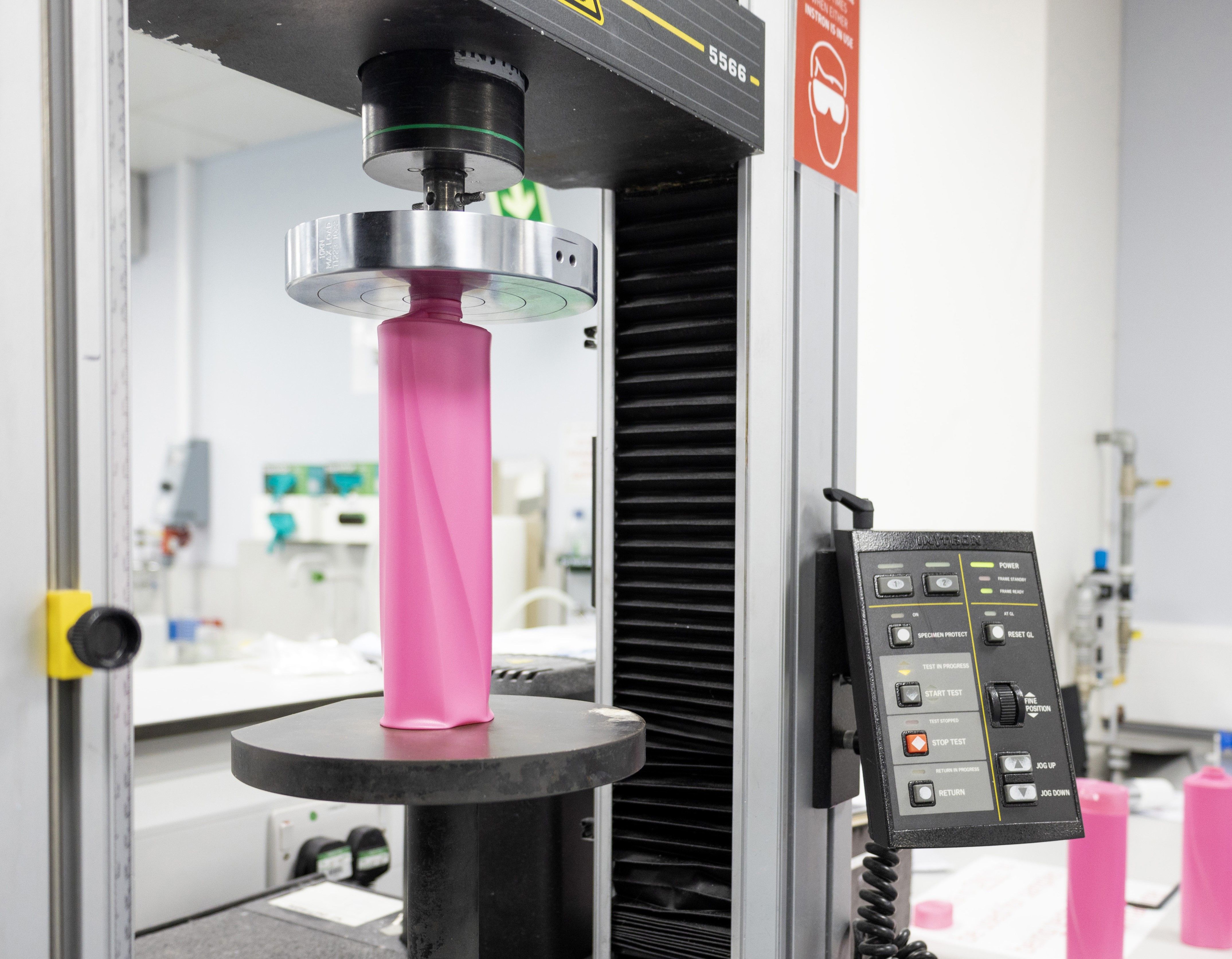 A close-up view of some lab testing equipment, showing a pink plastic bottle being compressed. The machine's control panel with various buttons is visible on the right.