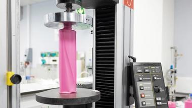 A close-up view of some lab testing equipment, showing a pink plastic bottle being compressed. The machine's control panel with various buttons is visible on the right.