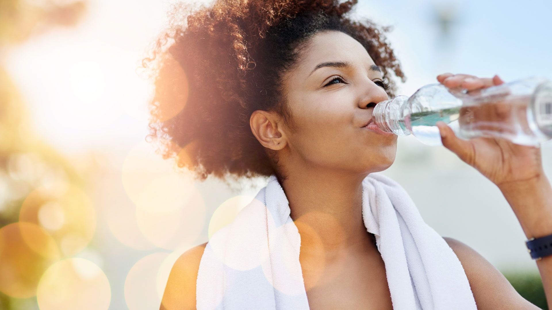 A person outdoors drinking from a clear water bottle, with a white towel draped around their shoulders and bright sunlight creating warm lens flares in the background.