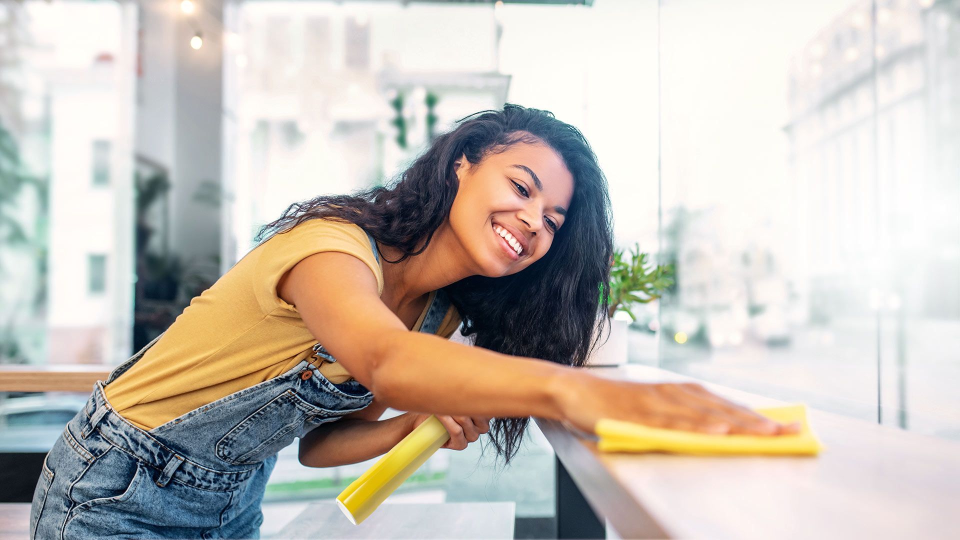 A young woman in overalls smiles while cleaning a surface with a yellow cloth inside a brightly lit room with large windows.